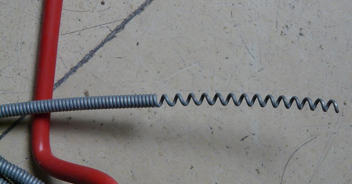 Coiled spring steel drain auger cable resting on a workbench beside a red handle