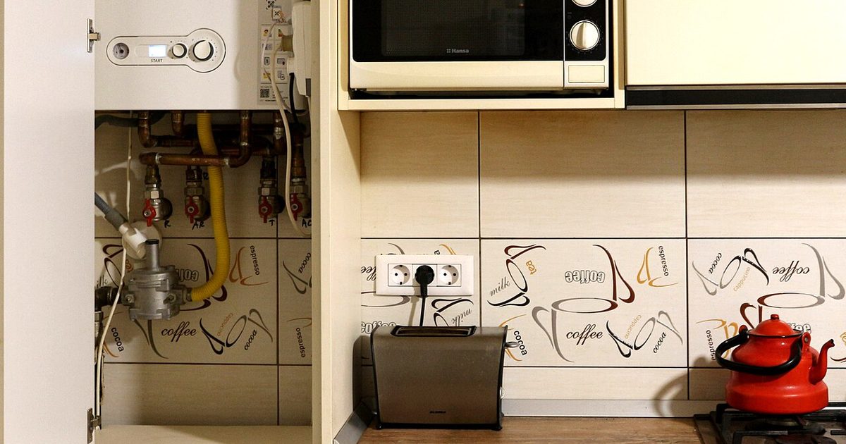 A wall-mounted gas water unit with copper pipework and a gas regulator inside a kitchen cupboard