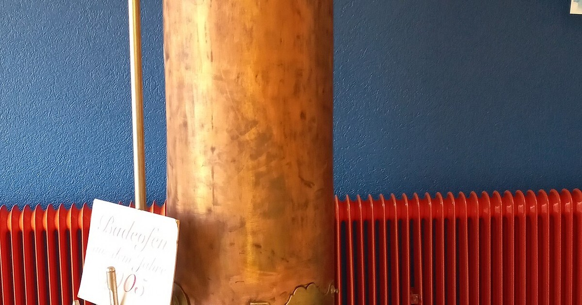 Copper hot water cylinder with red pipework connections against a wall